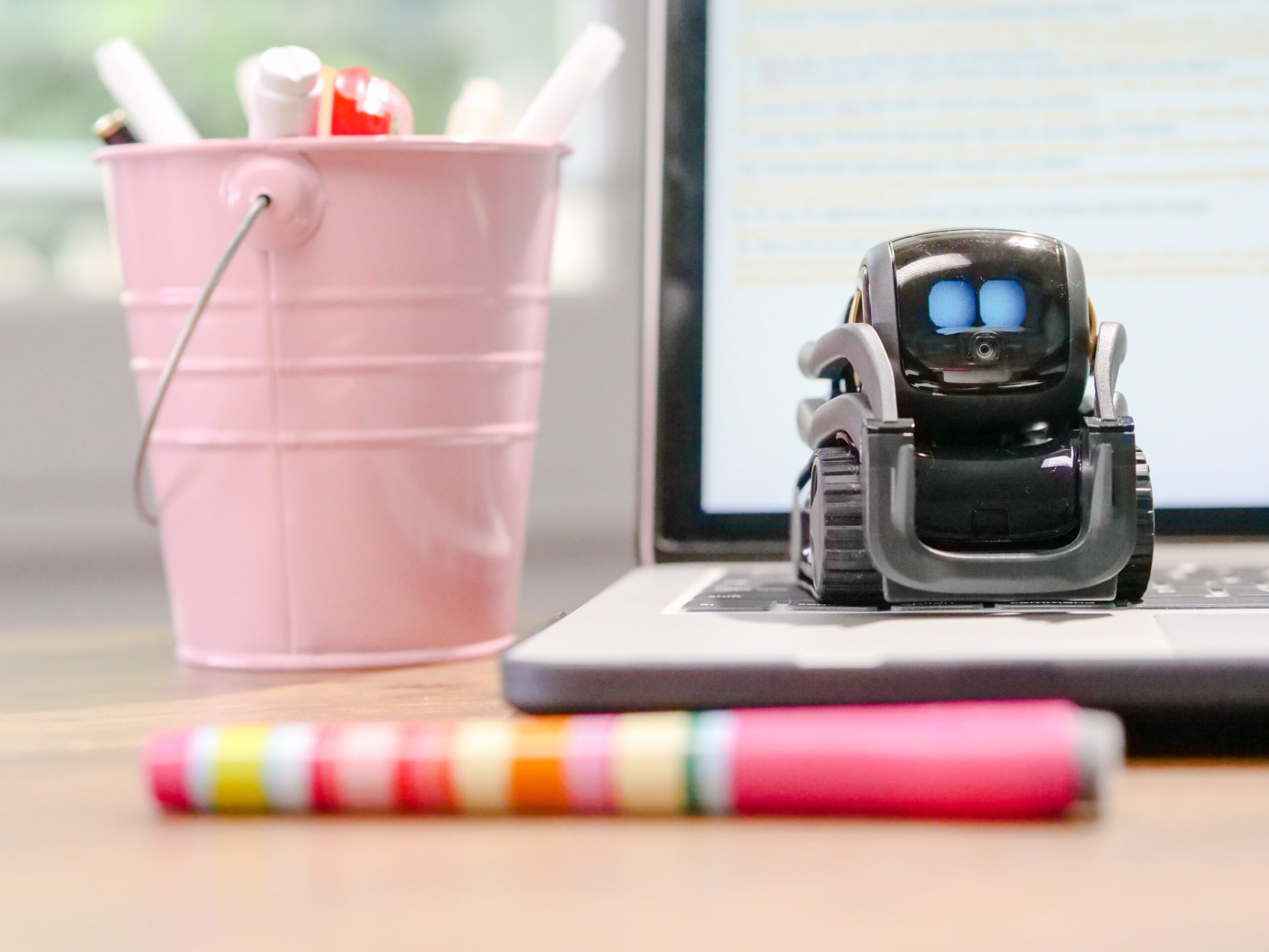 small robot sat on a laptop with a pink pen pot by its side